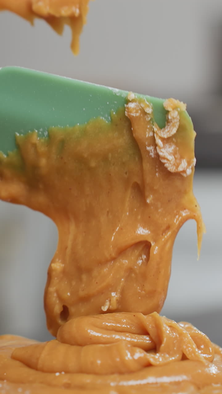 Close-up of thick, creamy brown batter falling from green spatula onto flour on countertop, smooth texture and rich consistency highlight homemade baking process
