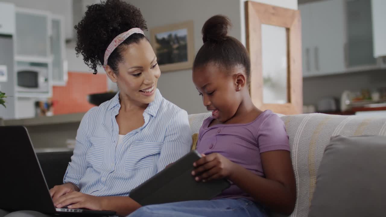 Happy african american mother and daughter on sofa, using laptop and tablet, slow motion, unaltered