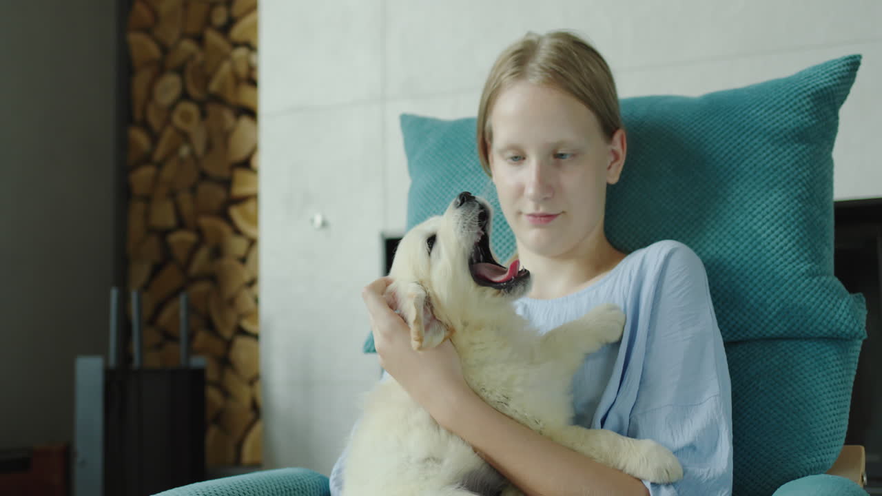 A teenage girl plays with a puppy of a golden retriever, sits in an armchair near the fireplace