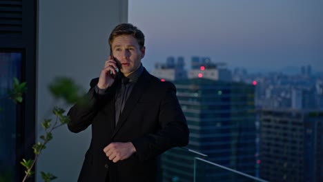 businessman in formal suit talking on smartphone at rooftop terrace during twilight in a modern cityscape - slow motion, parallax shot