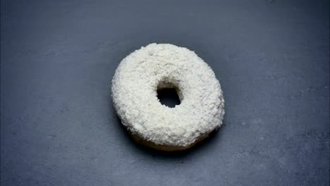 un primer plano de una rosquilla blanca con papas fritas de coco girando y mordidas en el fondo gris de la mesa.