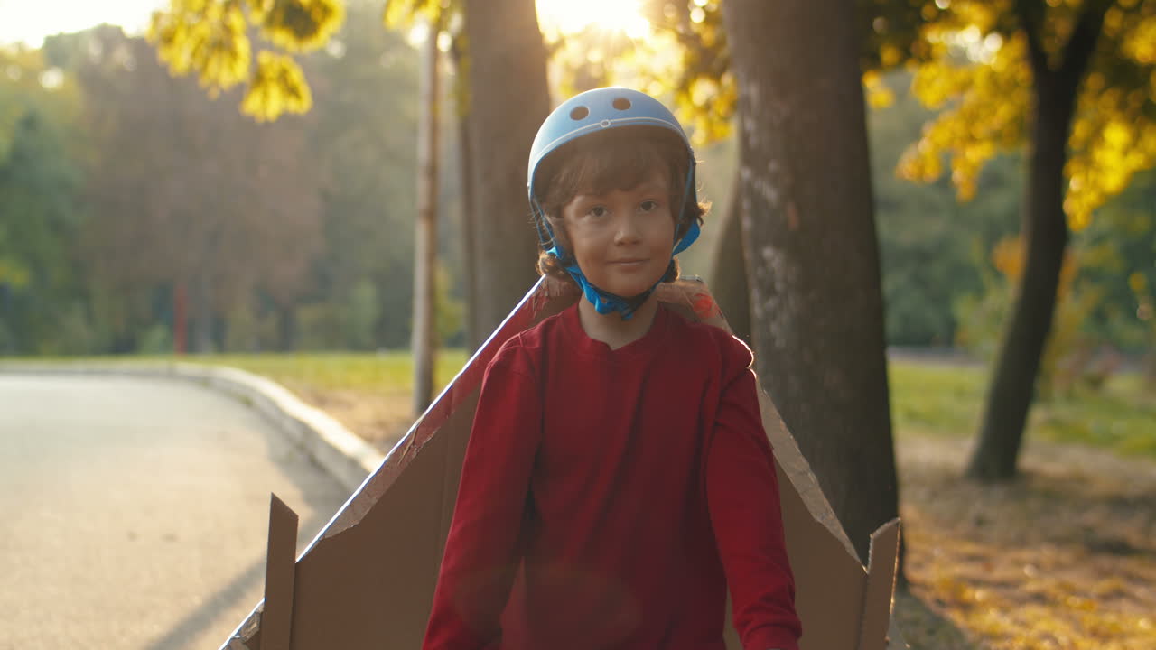 niño pequeño con casco y suéter rojo con alas de avión de cartón corriendo en el parque en un día soleado y jugando como piloto
