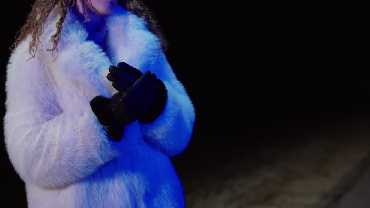 Ampia inquadratura cinematografica di una bella donna in piedi da sola in una strada tranquilla e vuota di notte, con un aspetto triste e infreddolito, che indossa un cappotto di pelliccia bianco e guanti di pelle. Concept di solitudine, crepacuore, freddo invernale