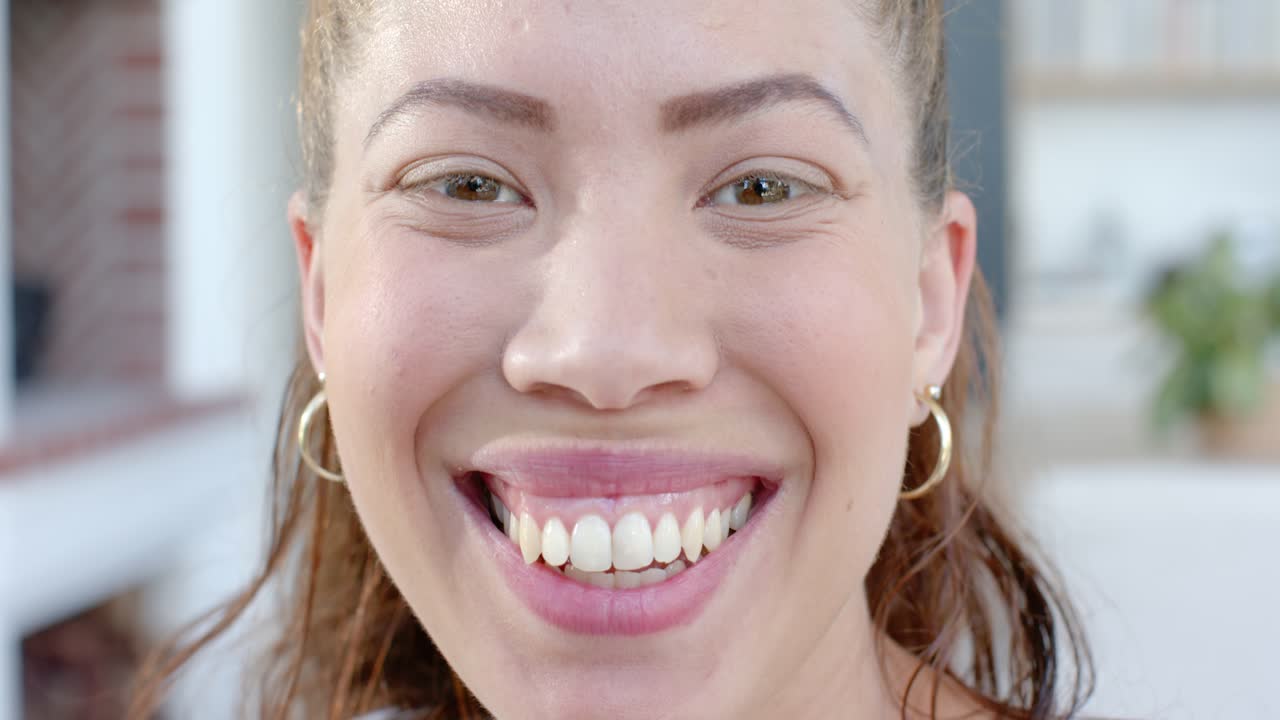 Mulher respondendo ao comando da câmera, sorrindo mais amplamente e inclinando-se para frente em casa, usando argolas de ouro.