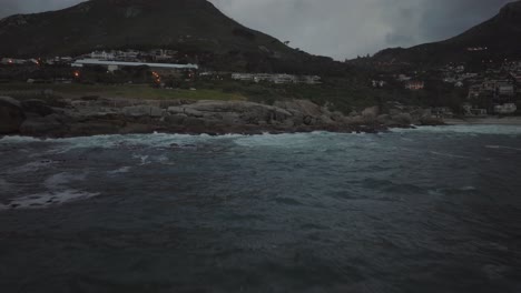 drone flying over the sea at camps bay beach in cape town south africa - it's cloudy and the waves are crashing against rocks with a view of the lion's head mountain