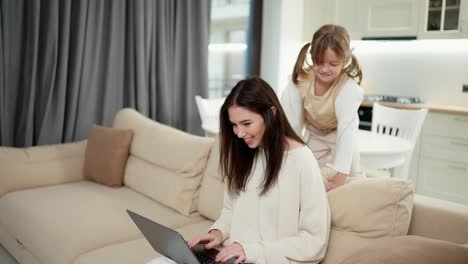 mother sit on sofa working on laptop computer distracted from daughter running play around
