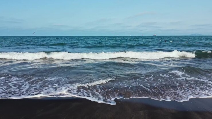 Onde si infrangono sulla riva sabbiosa in spiaggia a mezzogiorno con barche lontane nell'oceano.