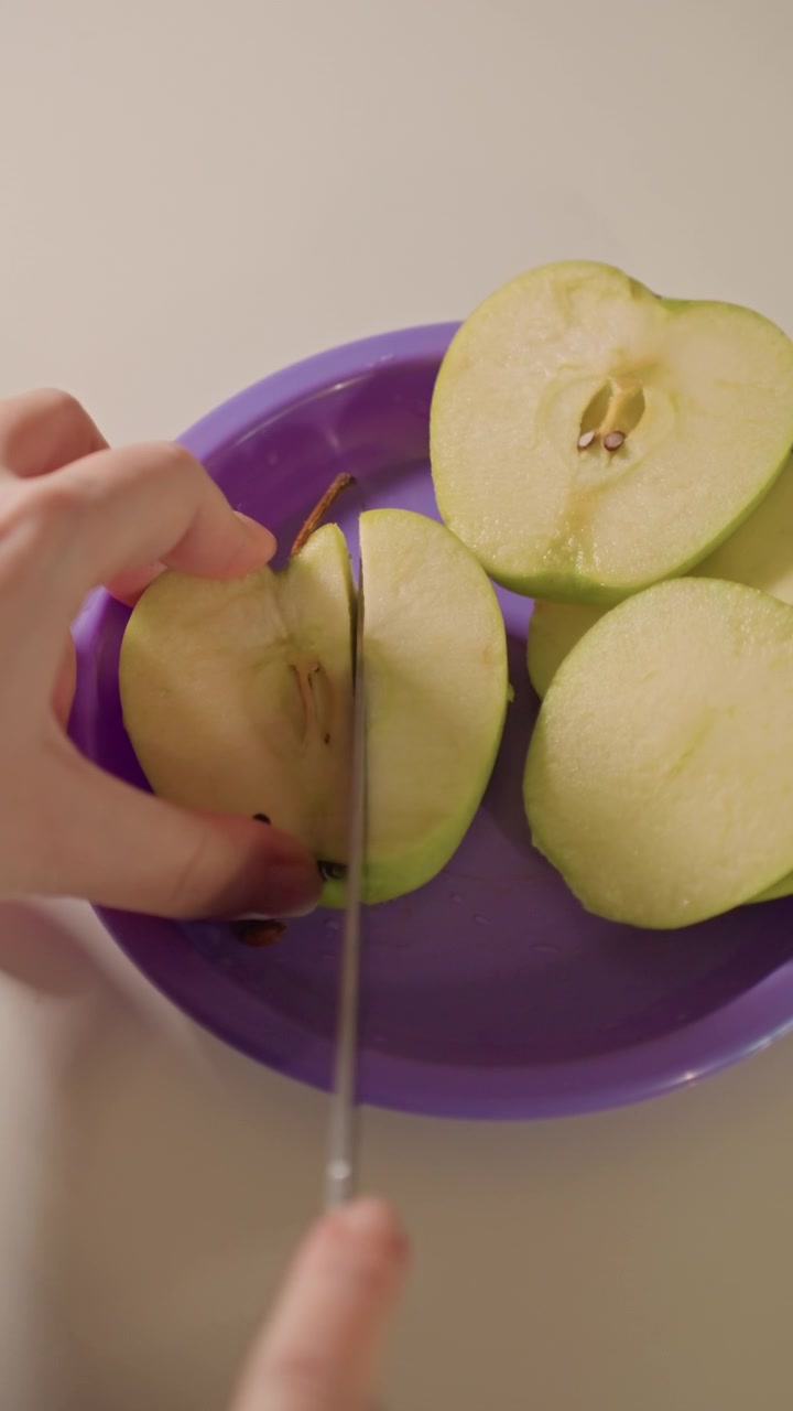 Primo piano di mani che affettano mele verdi su un piatto viola. Il coltello taglia le metà della mela, rivelando semi e dettagli del torsolo. Uno sfondo semplice enfatizza la preparazione del cibo.