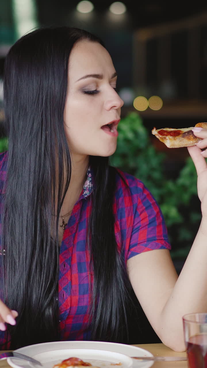 beautiful girl in a plaid shirt is eating a slice of pizza at a table in the pizzeria. Vertical video