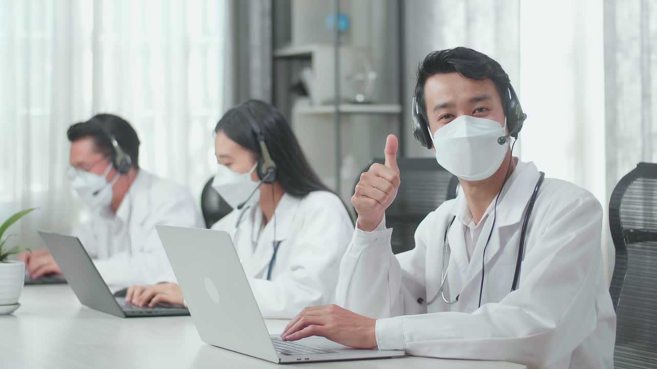A Man Of Three Asian Doctors With Stethoscope In Headset And Mask Working As Call Center Agent Look Up From Computer And Thumbs Up While His Colleagues Are Speaking And Typing During A Call At The Office.