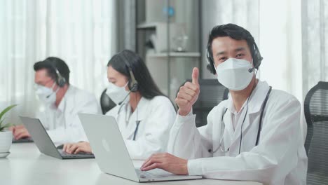 a man of three asian doctors with stethoscope in headset and mask working as call center agent look up from computer and thumbs up while his colleagues are speaking and typing during a call at the office.