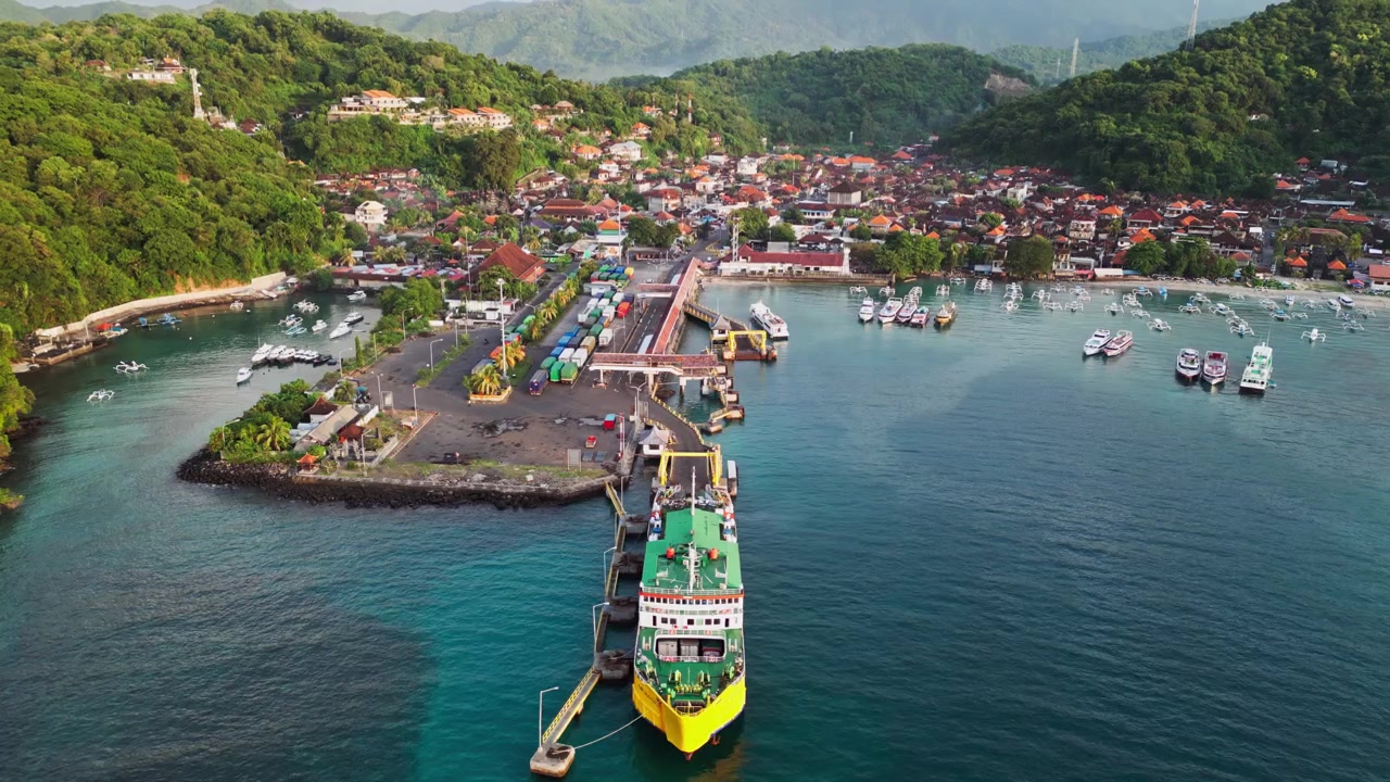 Porto di un'isola tropicale con traghetti passeggeri, imbarcazioni da diporto e un terminal dei traghetti accanto a una città costiera incorniciata da colline lussureggianti, riprese aeree con drone.