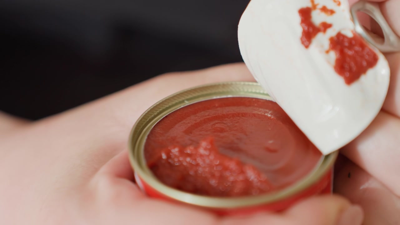 Primo piano di mani che aprono un piccolo contenitore di metallo staccando un coperchio di alluminio per rivelare una pasta rossa densa con granuli visibili, residui sulla parte inferiore del coperchio e un bordo lucido.