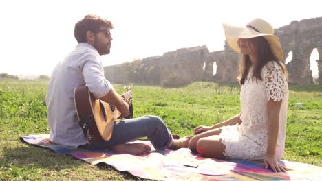 feliz joven pareja encantadora sentada acostada en una manta en la hierba frente a las ruinas del antiguo acueducto romano en el parco degli aquedotti parque romántico en roma tocar la guitarra cantar hermosa chica con sombrero bucólica cámara lenta