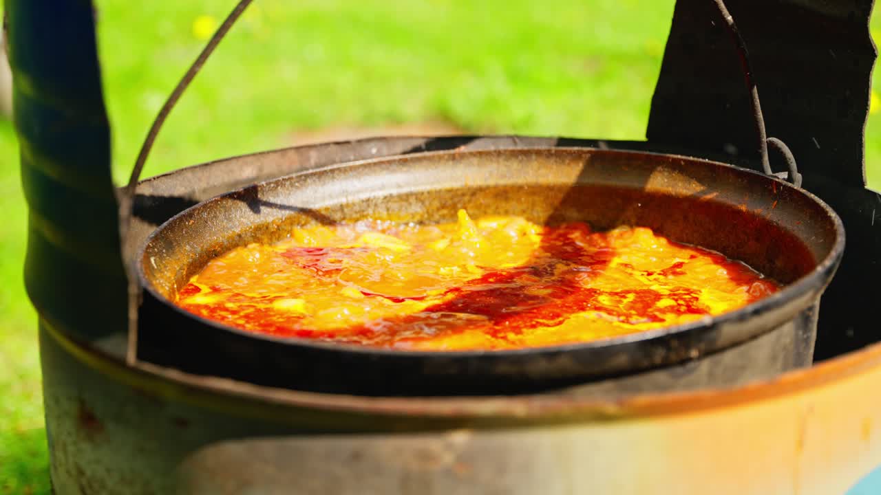 Bubbling soup simmers in black cauldron over open wood fire at rural outdoor site, slow motion