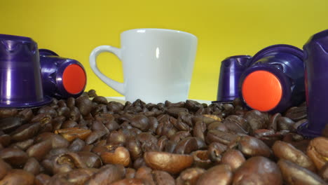 white mug with roasted coffee beans and plastic coffee capsule in front of yellow screen