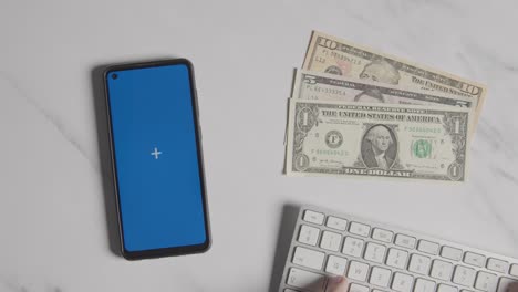 overhead currency shot of hand typing on computer keyboard with dollar notes and blue screen mobile phone