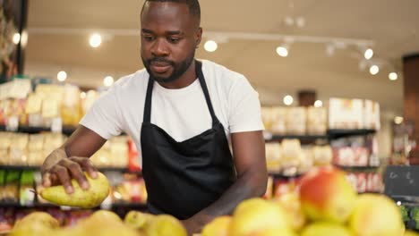Close-up:-A-black-man-in-a-white-T-shirt-and-black-apron-arranges-yellow-pears-on-a-supermarket-counter