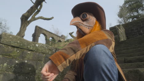 redhead wearing plague mask and cowboy hat sitting down with head turn towards camera