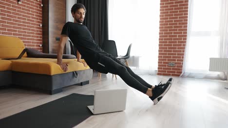 Man-athletic-doing-push-up-leaning-on-sofa-at-home-with-laptop