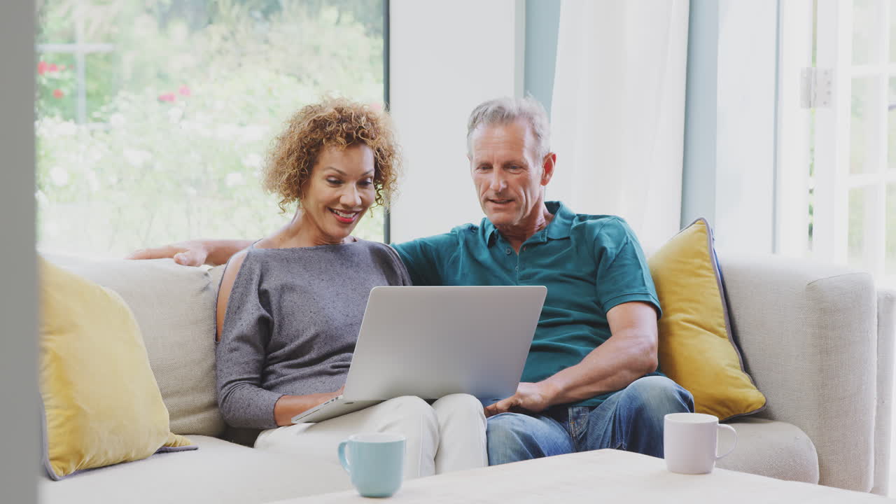 Senior Retired Couple Sitting On Sofa At Home Shopping Or Booking Holiday On Laptop