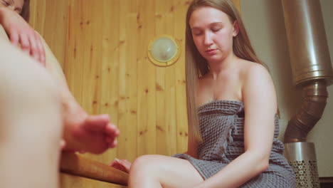 close up partial view of woman with tattoo seated beside younger lady applying pink scrub on leg with droplets falling onto wooden bench under steam in relaxing sauna skincare ritual