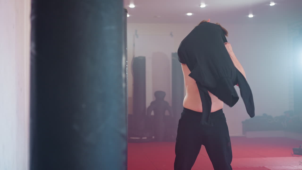 Cara se vestindo na academia, corpo sem camisa à mostra sob luzes fortes, segurando uma camisa preta com as duas mãos enquanto a veste, em pé no chão vermelho perto de sacos de pancada