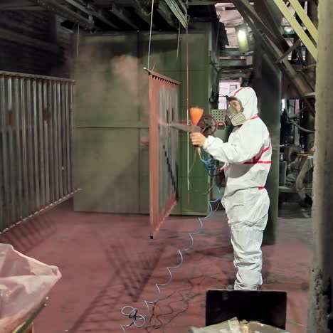 painting parts in small factory production. side view of a worker in protective suit working with a spray gun in a workshop. man paints metal structure with a pulverizer.