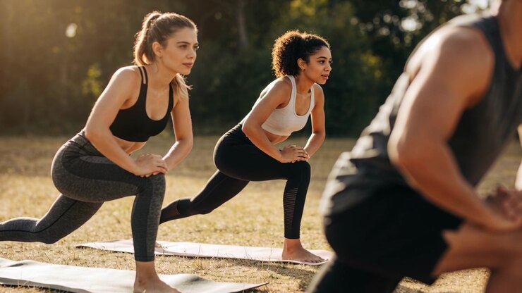 Responding to instructor cue, two women holding forward lunges on mats at sunny park, sportswear