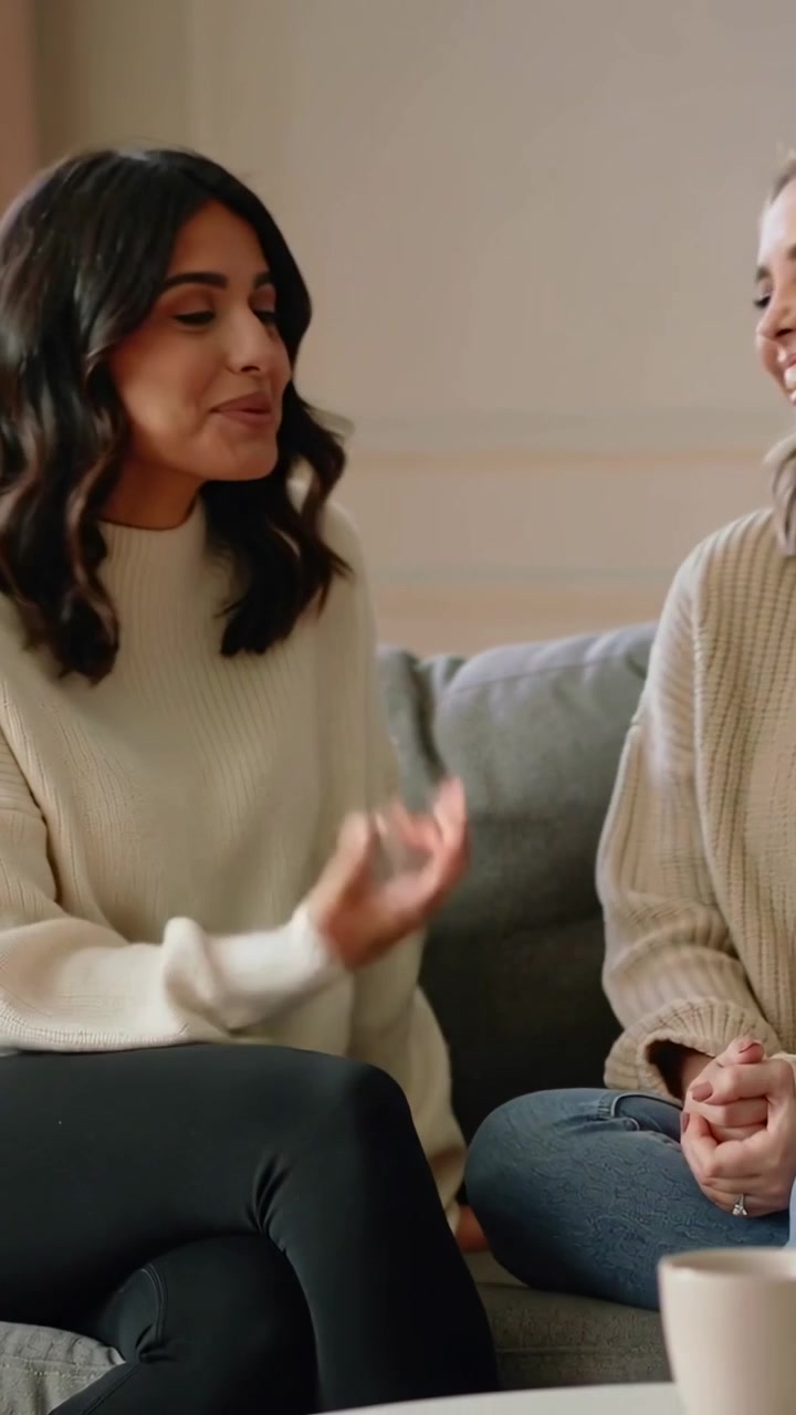 Vertikal video: Kvinde læner sig til venstre og starter en samtale, mens et par i trøjer og jeans griner på en grå sofa med et krus.