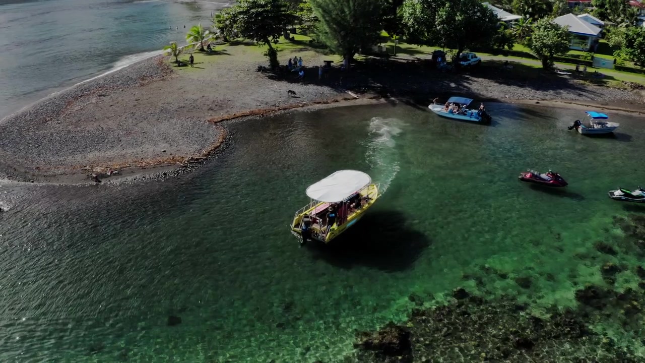 Drone che plana a bassa quota sopra barche da pesca allineate lungo la costa di sabbia nera a Teahupoo, Tahiti, con persone che si muovono tra gli scafi e l'acqua bassa