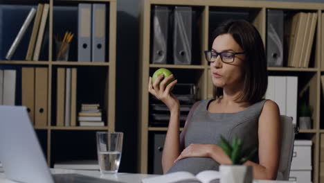 mujer embarazada con gafas descansando en su oficina en el trabajo, masticando una manzana y bebiendo agua de un vaso