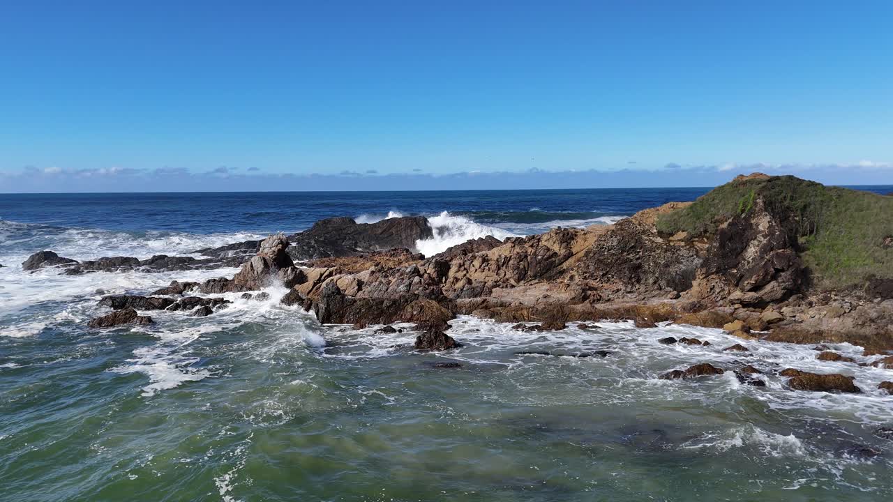 Drone footage captures waves crashing against Byron Bay's rugged coastline under clear blue skies