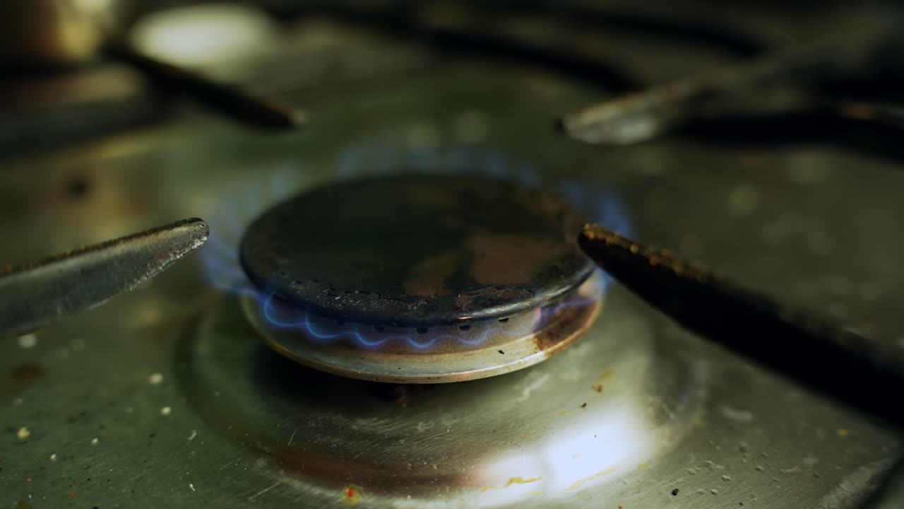 Primo piano di una fiamma blu del gas che si accende su un fornello sporco, a dimostrazione del calore della cottura domestica, del consumo energetico e del concetto di sicurezza.