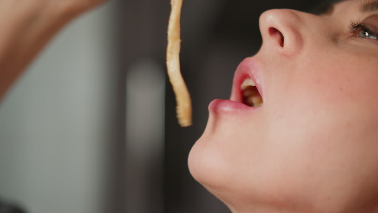 Close up upward view of young girl dragging long pasta into mouth using chopsticks with soft blur background, capturing intimate eating moment and texture of noodles