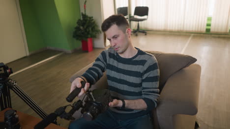 man reaching out with remote control, sitting on couch in living room, preparing video equipment. focused on setting up gear for filming or production. room features wood flooring, bright decor