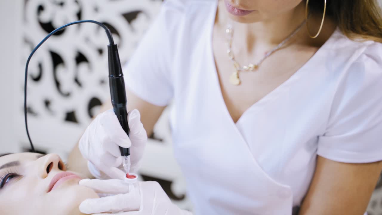 A woman in a white lab coat applies a pink pigment during the procedure of permanent lip makeup for a brunette. Close-up.