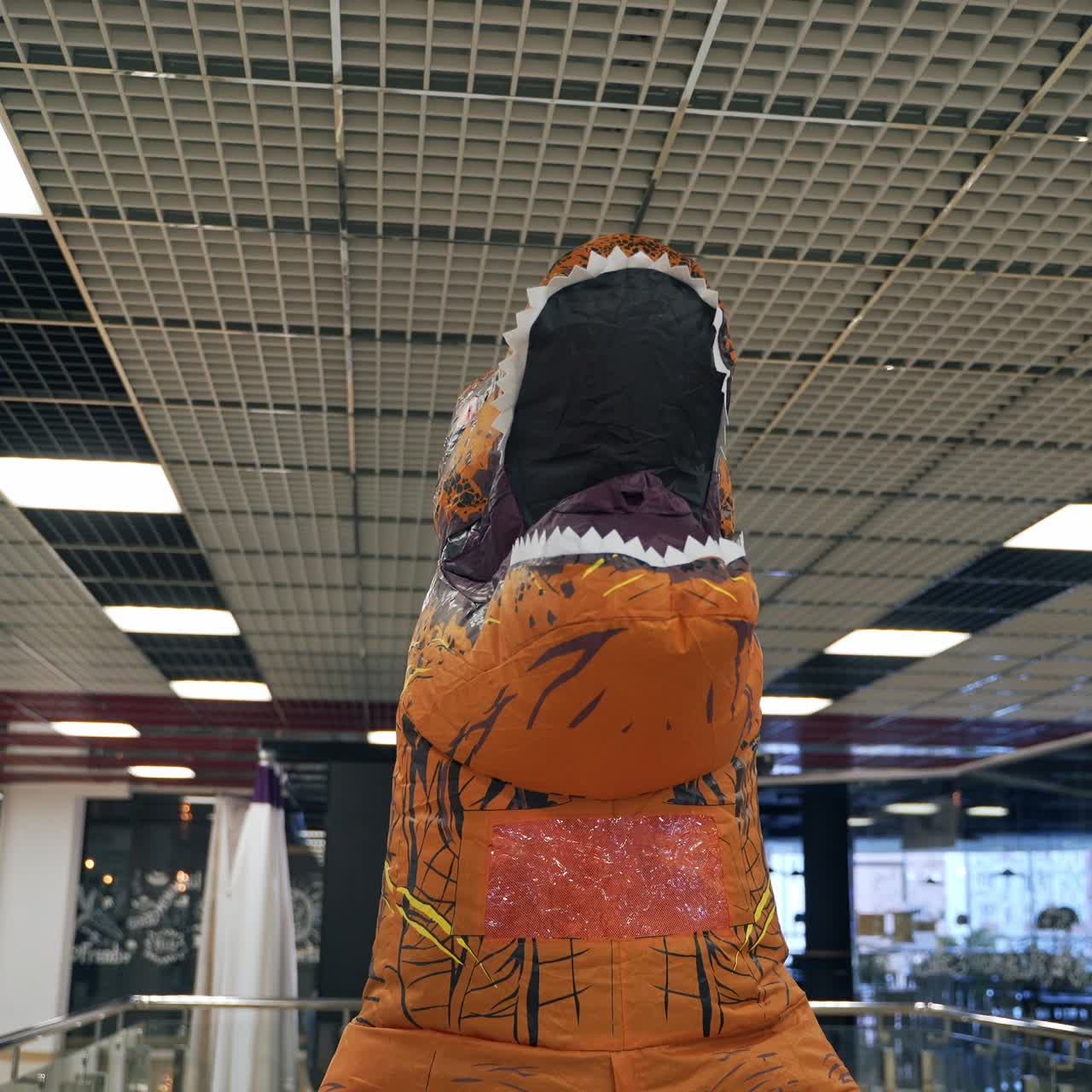 T-Rex dinosaur with opened mouth goes up by the escalator. Man in inflatable costume of orange dinosaur walking by the shopping mall