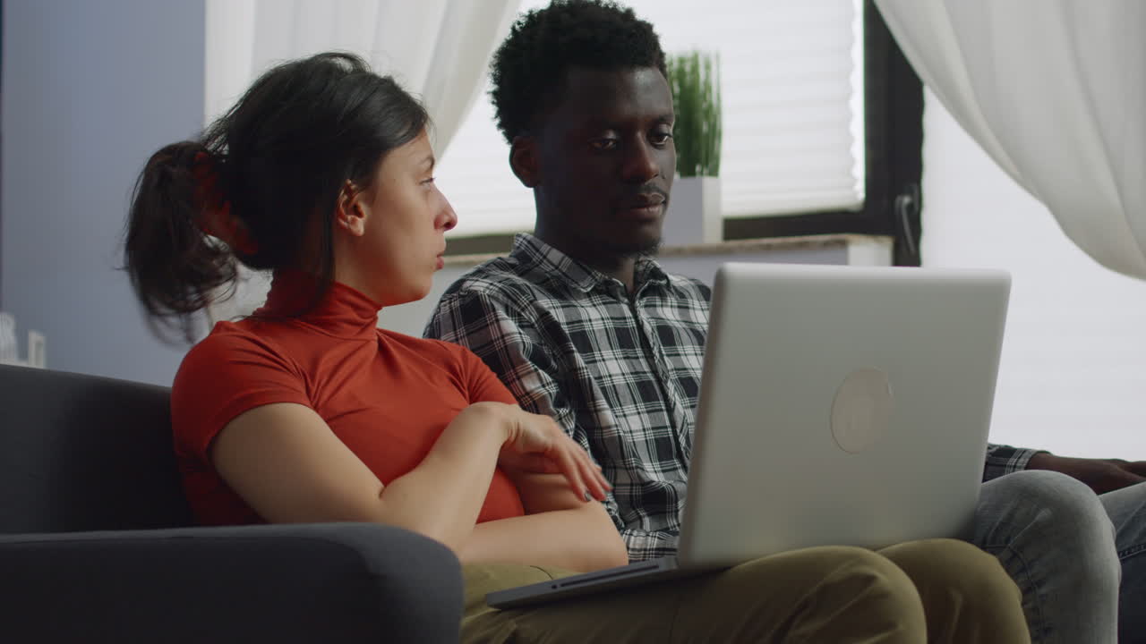 Interracial couple sitting on sofa using laptop in living room