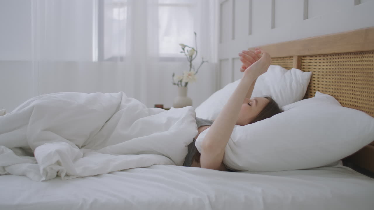 아름다운 갈색의 여자가 아침에 깨어나 침대에 누워 큰 창문에서  ⁇ 빛이 그녀를 비추고 행복한 젊은 소녀는 따뜻한  ⁇ 빛으로 새로운 날을 맞이합니다.