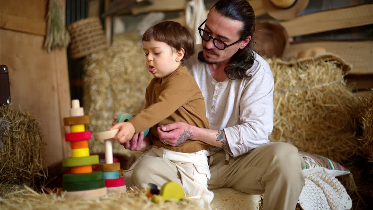 Father playing with his son with colourful, ecological wooden toys in a barn, near square hay bales