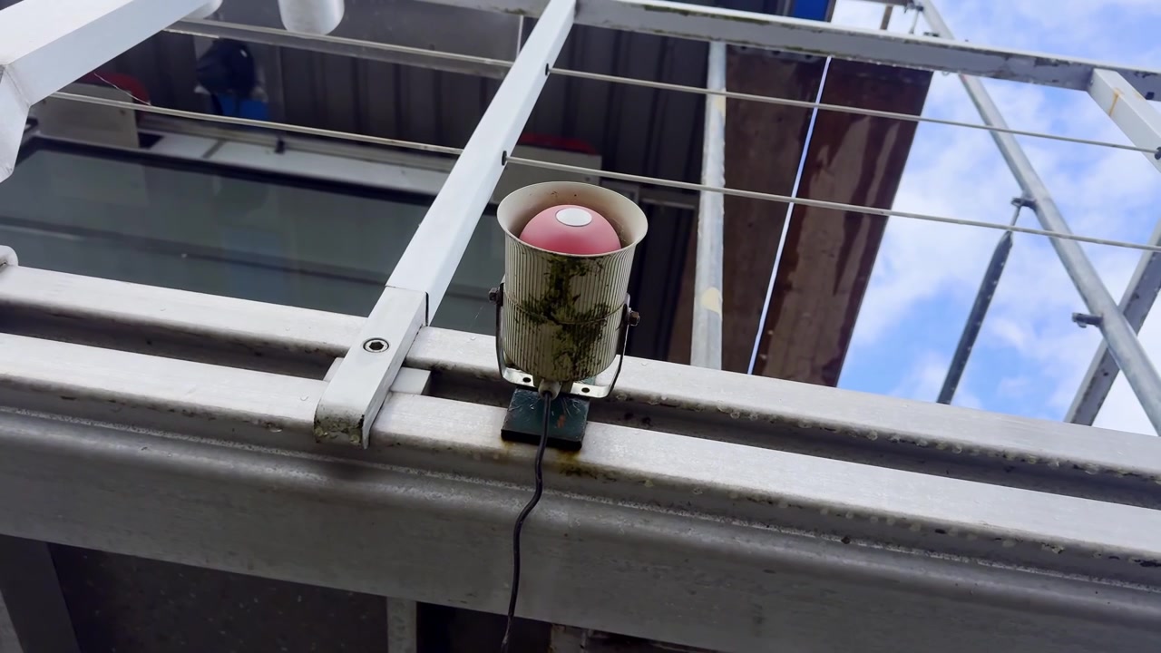 Primo piano di un altoparlante esterno o una sirena usurata montata sotto una struttura metallica, con ringhiere, cavi, macchie e riflessi del cielo in un dettaglio di infrastruttura urbana
