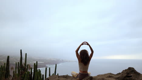 Against-the-backdrop-of-the-ocean-and-green-mountains,-a-woman-meditates-atop-a-mountain,-her-hands-forming-the-Maha-Sakal-gesture