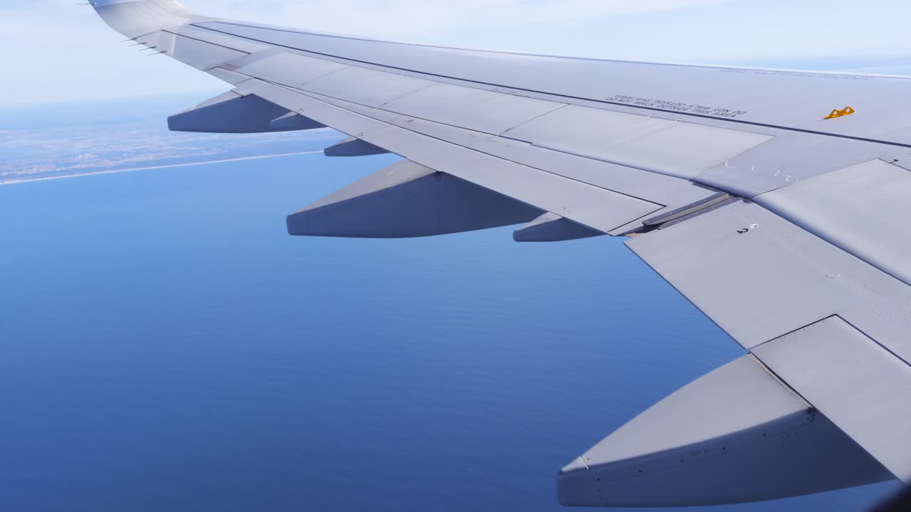 Solrik utsikt fra et passasjervindu av en flyvinge som glir over dypt, blått Atlanterhavsvann, med Portugals kystlinje og strender synlig i det fjerne