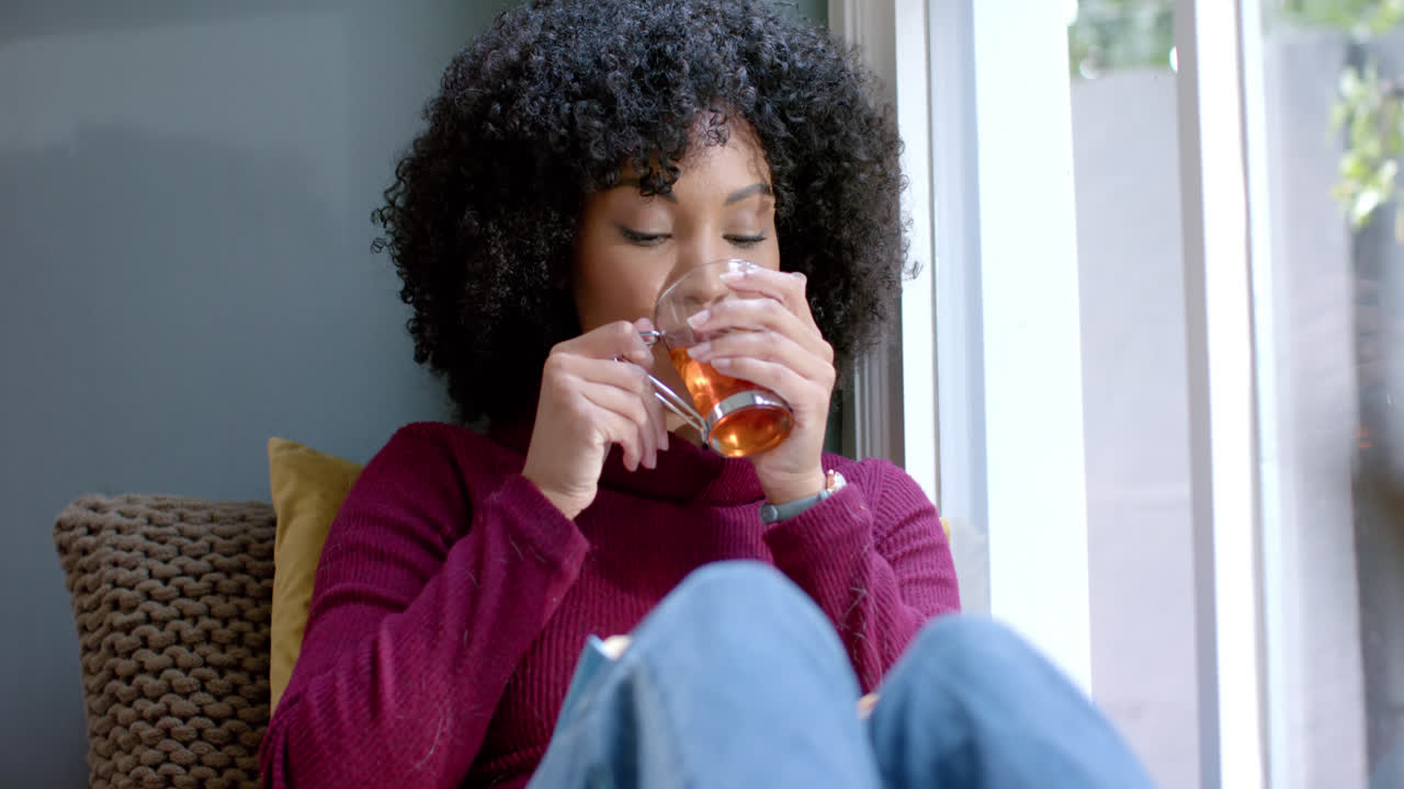 Happy biracial woman drinking tea and reading book at home, slow motion