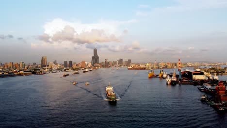 Ein-Geschäftiger-Hafen-Mit-Schiffen-Und-Kränen,-Die-Skyline-Der-Stadt-Im-Hintergrund,-Goldene-Stunde,-Luftaufnahme