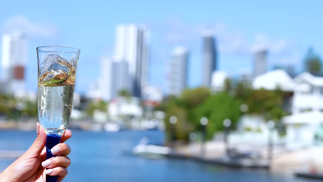 A hand gently swirls a champagne glass, creating a dynamic liquid motion against a sunlit, blurred city waterfront backdrop. Bright daylight enhances the celebratory mood