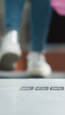 vista trasera de una mujer joven caminando hacia la escalera mecánica en zapatillas deportivas y vaqueros casuales, con una bolsa de compras borrosa y un moderno centro comercial interior en el fondo
