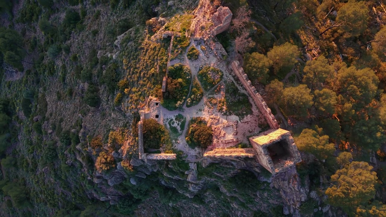 Rovine di un castello storico arroccate su una collina rocciosa nella Sierra Calderona, Spagna, circondate da una fitta foresta bagnata dalla calda luce dorata del tramonto.