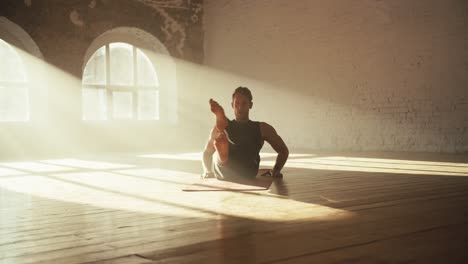 a man in a black sports summer uniform is doing a scissors exercise in a sunny brick hall. fitness classes on a special mat on